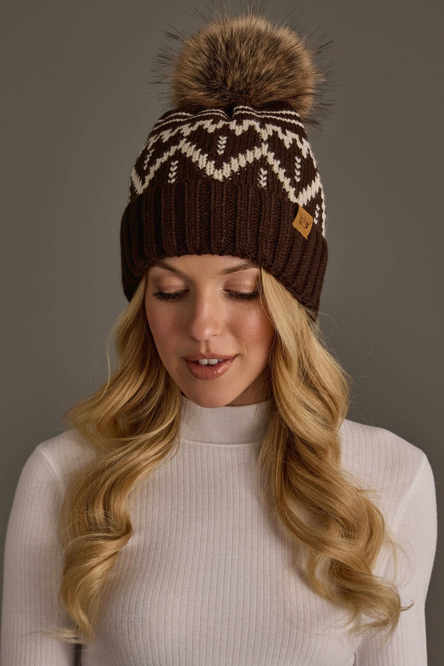 A woman with long blonde hair wears the Chocolate Brown & Beige Patterned Pom Hat from Panache Apparel Collection, featuring a white design. She is looking down and wearing a white ribbed turtleneck sweater.