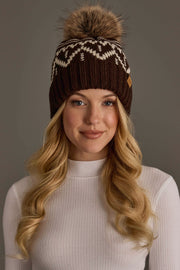 A woman with long blonde hair wears the Panache Apparel Collection’s Chocolate Brown & Beige Patterned Pom Hat, a brown knit hat featuring white patterns and a large faux fur pom-pom, paired with a white ribbed turtleneck, as she smiles gently at the camera against a plain background.