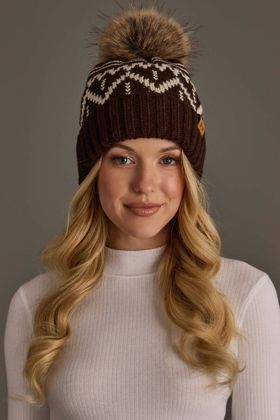 A woman with long blonde hair wears the Panache Apparel Collection’s Chocolate Brown & Beige Patterned Pom Hat, a brown knit hat featuring white patterns and a large faux fur pom-pom, paired with a white ribbed turtleneck, as she smiles gently at the camera against a plain background.