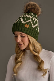A woman with long, curled blonde hair looks down and smiles slightly while wearing the Olive & Beige Patterned Pom Hat by Panache Apparel Collection, a green knit hat featuring white patterns and a large brown pom-pom, paired with a white ribbed sweater.