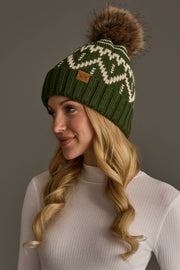 A woman with long blonde curls wears the Panache Apparel Collection Olive & Beige Patterned Pom Hat, featuring a white mountain design and brown pom-pom, along with a white ribbed top, against a plain gray background.