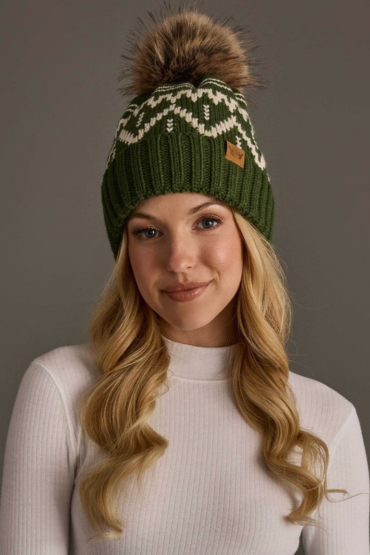 A woman with long blonde hair wears a white turtleneck and the Olive & Beige Patterned Pom Hat from Panache Apparel Collection, featuring a green knit design with a white pattern and a large fur pom-pom, standing against a plain gray background.
