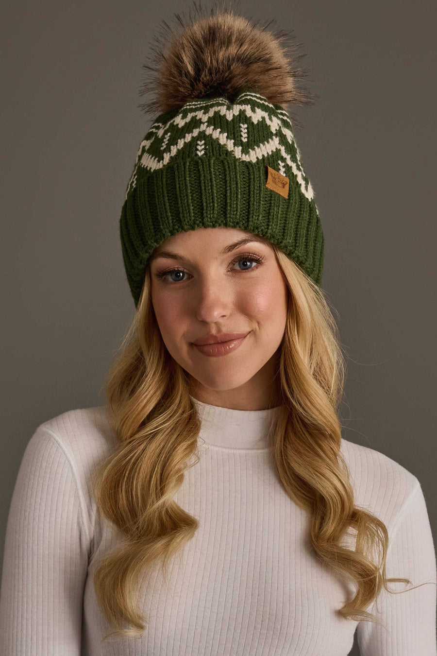 A woman with long blonde hair wears a white turtleneck and the Olive & Beige Patterned Pom Hat from Panache Apparel Collection, featuring a green knit design with a white pattern and a large fur pom-pom, standing against a plain gray background.