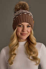 A woman with long blonde hair smiles at the camera against a plain gray background, wearing the Panache Apparel Collection’s Mocha, Light Taupe & Brown Patterned Pom Hat and a white ribbed top.
