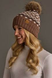 A woman with long blonde hair, smiling and looking to the side, wears the Panache Apparel Collection’s Mocha, Light Taupe & Brown Patterned Pom Hat, a knit beanie topped with a fur pom-pom, paired with a white ribbed turtleneck top against a neutral background.
