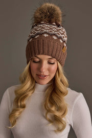 A woman with long blonde hair is wearing the Panache Apparel Collection’s Mocha, Light Taupe & Brown Patterned Pom Hat, a brown knit winter hat featuring a fur pom-pom and white patterned design, paired with a white ribbed turtleneck sweater, as she looks down with a neutral expression.