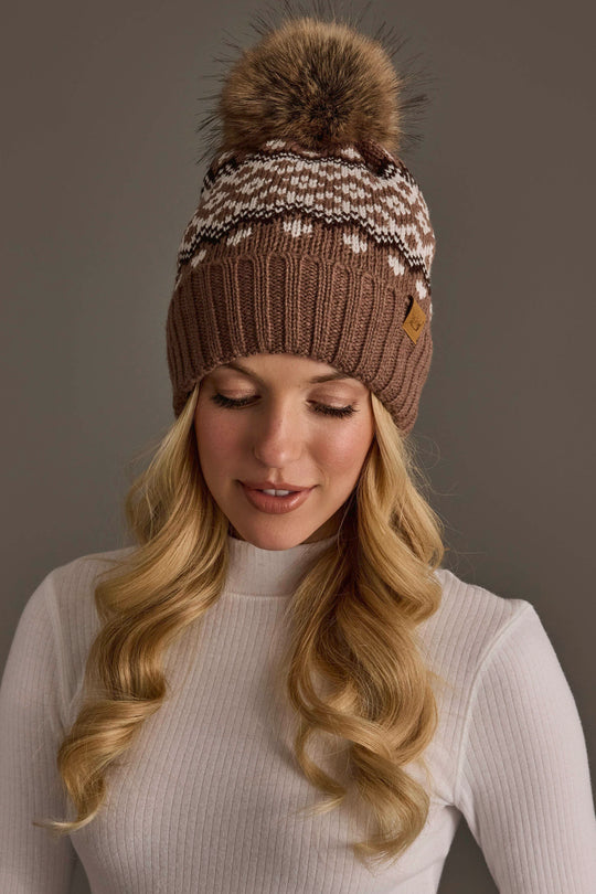 A woman with long blonde hair is wearing the Panache Apparel Collection’s Mocha, Light Taupe & Brown Patterned Pom Hat, a brown knit winter hat featuring a fur pom-pom and white patterned design, paired with a white ribbed turtleneck sweater, as she looks down with a neutral expression.