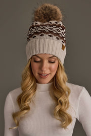 A woman with long blonde hair wears a white knit sweater and the Panache Apparel Collection’s Light Taupe, Brown & Mocha Patterned Pom Hat, featuring a brown design and faux fur pom-pom, as she looks down with a soft smile against a gray background.