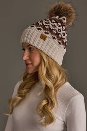 A woman with long blonde hair wears the Light Taupe, Brown & Mocha Patterned Pom Hat from Panache Apparel Collection along with a light ribbed turtleneck sweater, against a plain gray background.