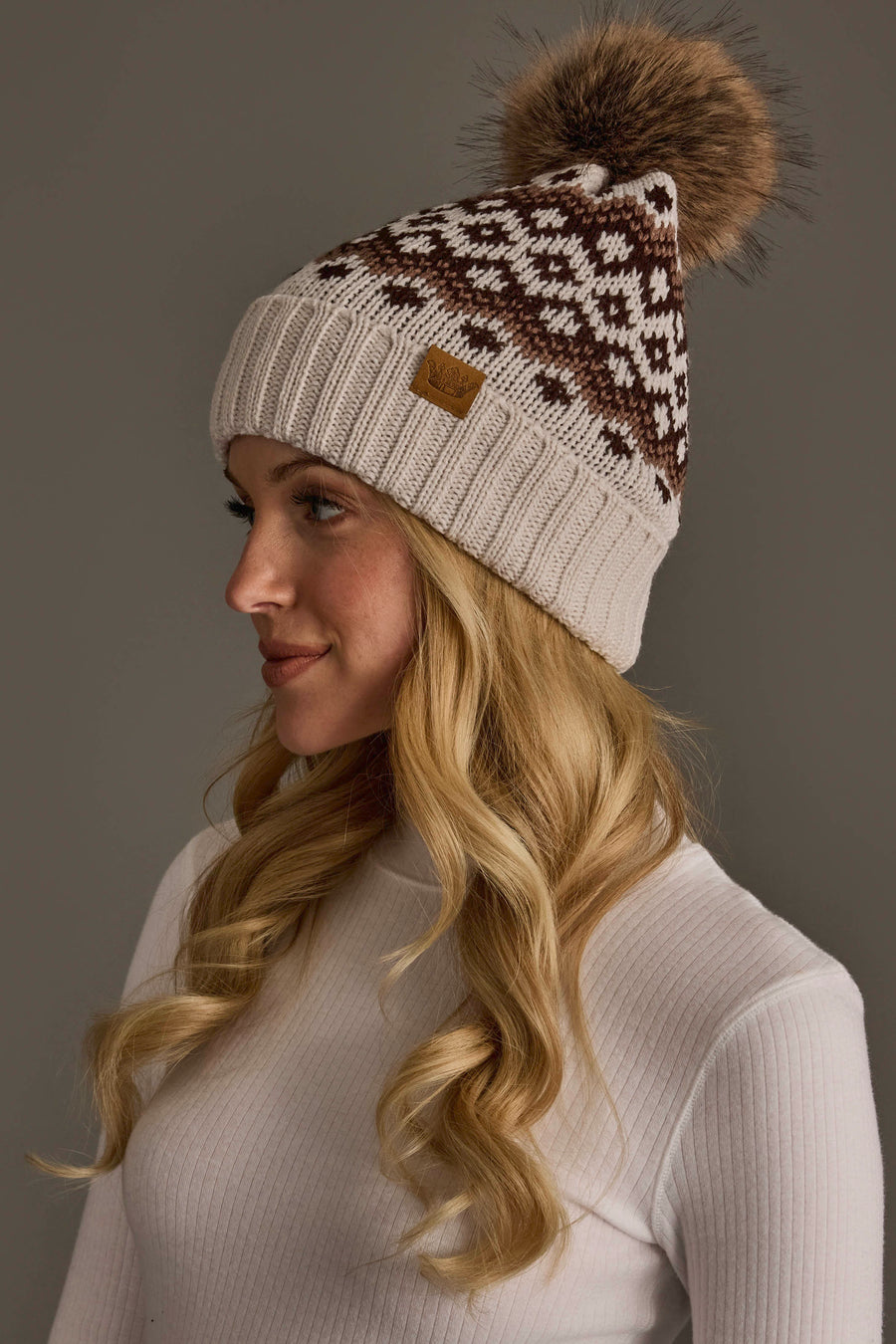 A woman with long blonde hair wears the Light Taupe, Brown & Mocha Patterned Pom Hat from Panache Apparel Collection along with a light ribbed turtleneck sweater, against a plain gray background.