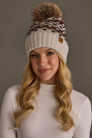 A woman with long blonde hair wearing a white ribbed sweater and the Panache Apparel Collection Light Taupe, Brown & Mocha Patterned Pom Hat, featuring a brown and white knit design and a large faux fur pom-pom, stands against a plain gray background.