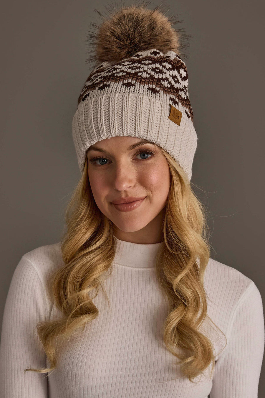 A woman with long blonde hair wearing a white ribbed sweater and the Panache Apparel Collection Light Taupe, Brown & Mocha Patterned Pom Hat, featuring a brown and white knit design and a large faux fur pom-pom, stands against a plain gray background.