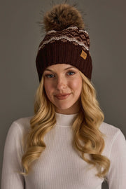 A woman with long blonde hair models the Panache Apparel Collection Brown, Mocha & Light Taupe Patterned Pom Hat, featuring a knit design with a pom-pom and white and gray accents, paired with a white ribbed long-sleeve top against a plain gray background.