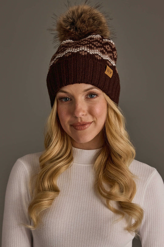 A woman with long blonde hair models the Panache Apparel Collection Brown, Mocha & Light Taupe Patterned Pom Hat, featuring a knit design with a pom-pom and white and gray accents, paired with a white ribbed long-sleeve top against a plain gray background.