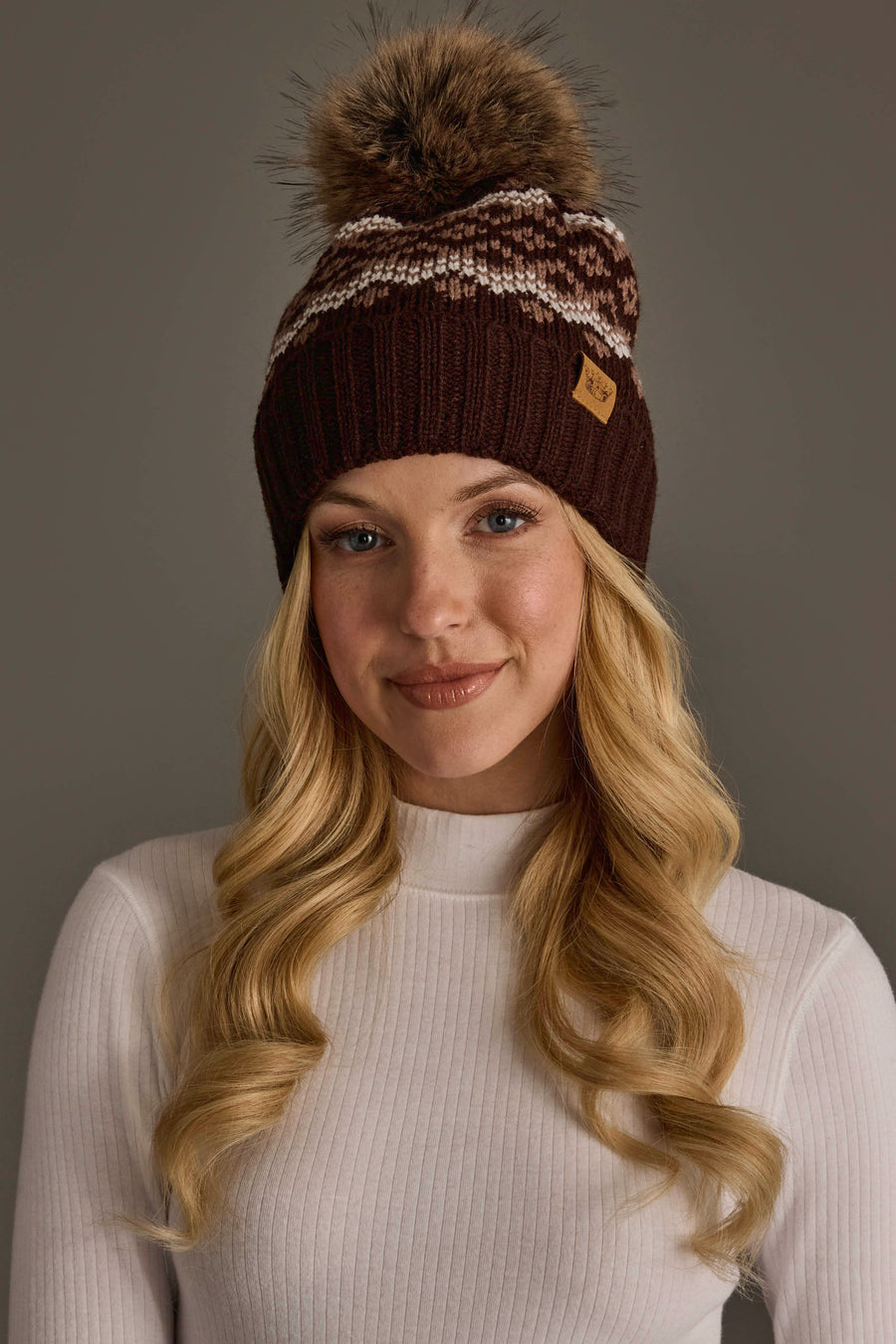 A woman with long blonde hair models the Panache Apparel Collection Brown, Mocha & Light Taupe Patterned Pom Hat, featuring a knit design with a pom-pom and white and gray accents, paired with a white ribbed long-sleeve top against a plain gray background.