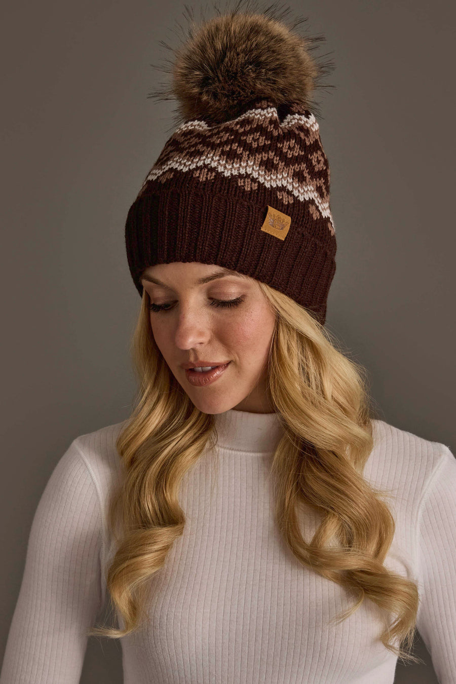 A woman with long blonde hair looks down and smiles softly in front of a plain gray background, wearing the Panache Apparel Collection Brown, Mocha & Light Taupe Patterned Pom Hat along with a white ribbed long-sleeve shirt.
