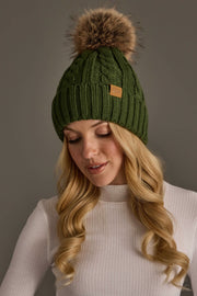 A woman with long blonde hair wears the Panache Apparel Collection’s Dark Green Cable Knit Design Pom Hat, featuring a brown faux fur pom-pom, paired with a white ribbed turtleneck sweater as she looks down against a neutral background.