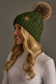 A woman with long blonde hair, wearing the Panache Apparel Collection Dark Green Cable Knit Design Pom Hat featuring a large brown faux fur pom-pom and a white ribbed turtleneck, smiles while facing left against a plain background.