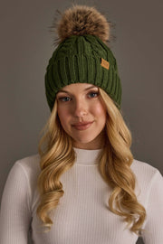 A woman with long blonde hair wears the Panache Apparel Collection’s Dark Green Cable Knit Design Pom Hat, featuring a brown faux fur pom-pom, paired with a white ribbed turtleneck sweater, as she stands against a plain gray background.