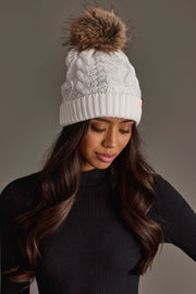 A woman with long wavy brown hair is wearing the Panache Apparel Collection White Cable Knit Pom Hat and a black ribbed top, looking down against a neutral background.