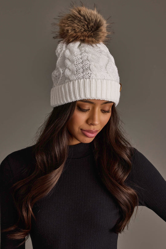 A woman with long wavy brown hair is wearing the Panache Apparel Collection White Cable Knit Pom Hat and a black ribbed top, looking down against a neutral background.