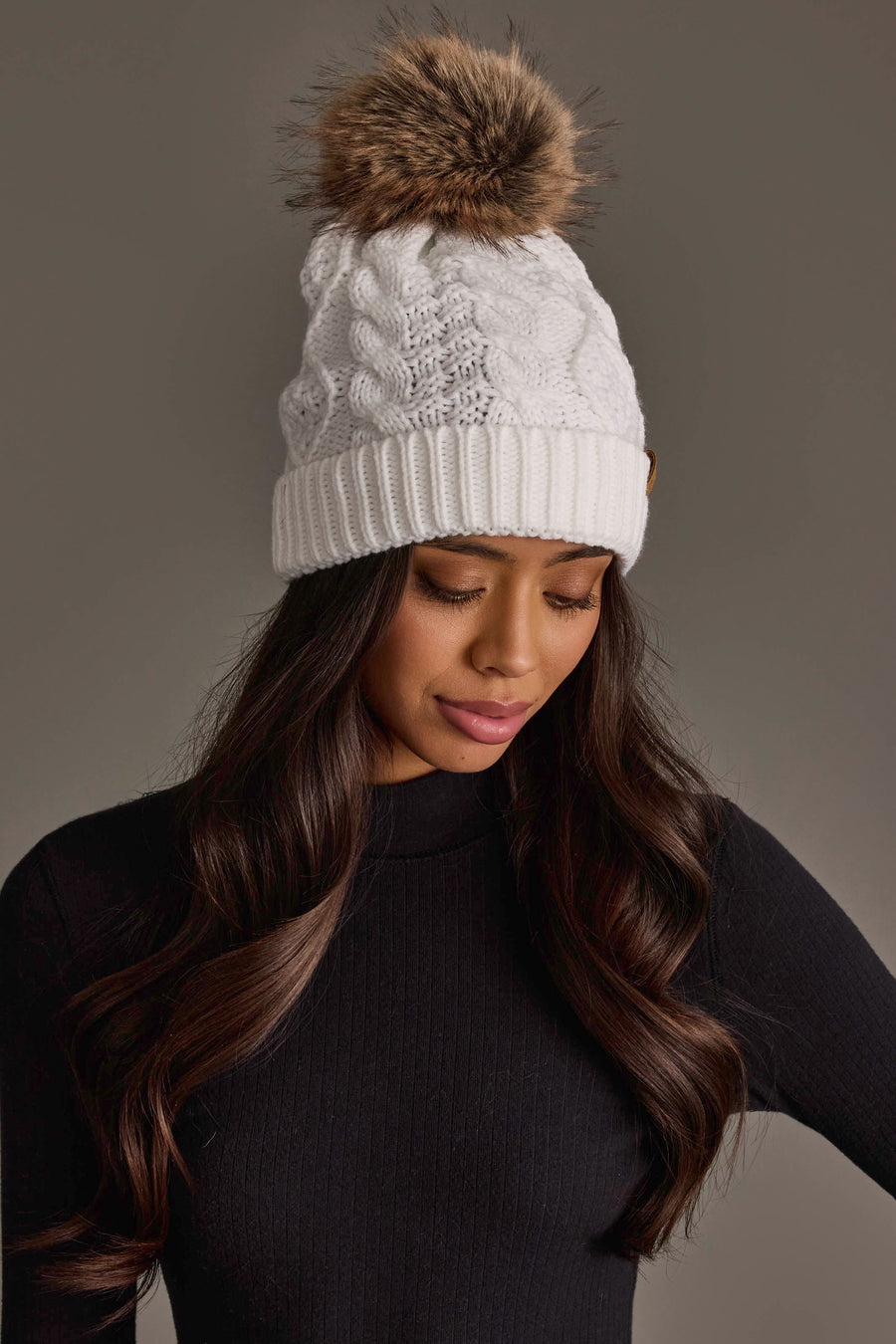 A woman with long wavy brown hair is wearing the Panache Apparel Collection White Cable Knit Pom Hat and a black ribbed top, looking down against a neutral background.