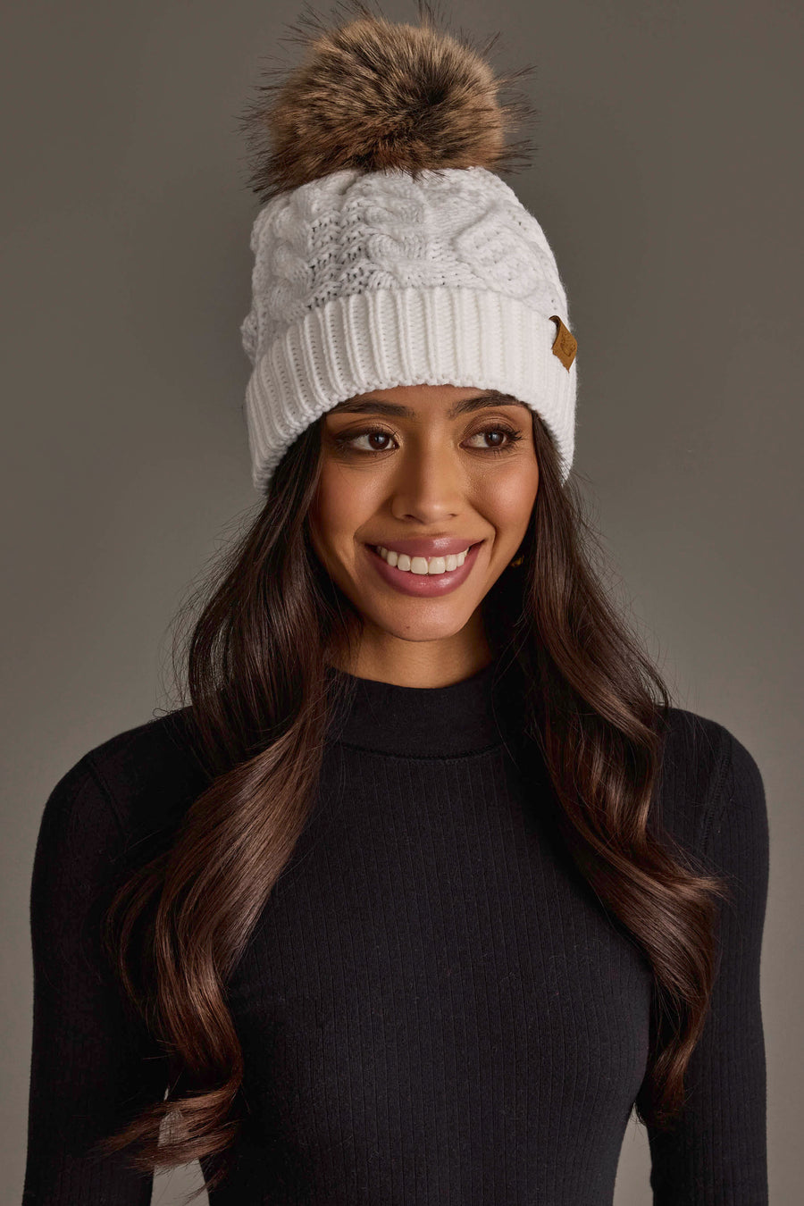 A woman with long dark hair is smiling against a plain gray background, wearing the Panache Apparel Collection White Cable Knit Pom Hat, featuring a faux fur pom-pom and brown tag, and a black ribbed sweater.