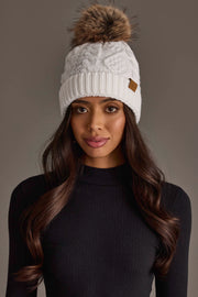 A woman with long brown hair is wearing the Panache Apparel Collection’s White Cable Knit Pom Hat and a black ribbed turtleneck sweater, standing in front of a plain gray background.