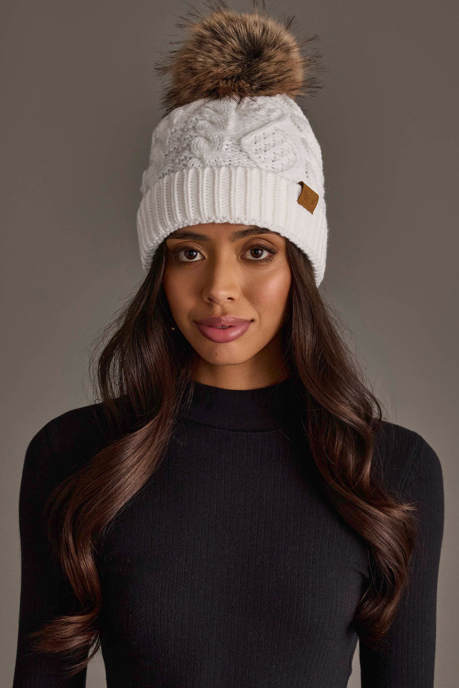 A woman with long brown hair is wearing the Panache Apparel Collection’s White Cable Knit Pom Hat and a black ribbed turtleneck sweater, standing in front of a plain gray background.