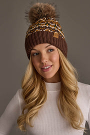 A woman with long blonde hair wears the Panache Apparel Collection Brown, Mustard & Cream Patterned Pom Hat and a white ribbed sweater, smiling and gazing slightly to the side against a plain background.