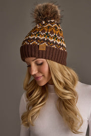 A woman with long blonde hair wears the Brown, Mustard & Cream Patterned Pom Hat by Panache Apparel Collection and a white ribbed turtleneck sweater, looking down and smiling softly against a plain background.