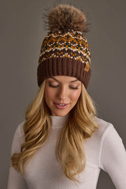 A woman with long blonde hair wears the Panache Apparel Collection’s Brown, Mustard & Cream Patterned Pom Hat, styled with a white ribbed turtleneck and posing against a plain gray background.