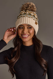 A woman wearing a black long-sleeve top and the Panache Apparel Collection Beige, Mustard & Green Patterned Pom Hat smiles and touches the side of her hat, standing against a plain gray background.
