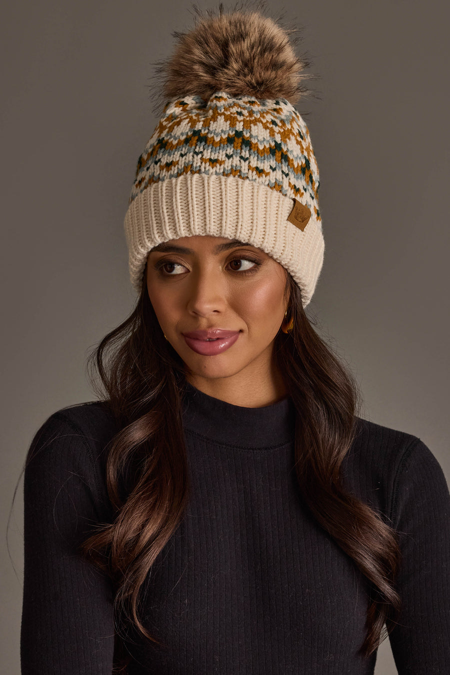 Model wearing the Panache Apparel Collection’s Beige, Mustard & Green Patterned Pom Hat, a knit beanie featuring a geometric design and a fur pom-pom, paired with a black ribbed turtleneck, gazing slightly to the side against a plain gray background.