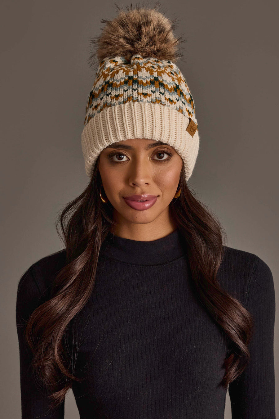 A woman with long, wavy dark hair wears the Panache Apparel Collection’s Beige, Mustard & Green Patterned Pom Hat and a black ribbed sweater, standing against a neutral background.