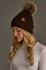 A woman with long, wavy blonde hair wears the Chocolate Brown Knit Pom Hat from Panache Apparel Collection, featuring a fur pom-pom, and a white ribbed turtleneck as she looks down against a neutral background.