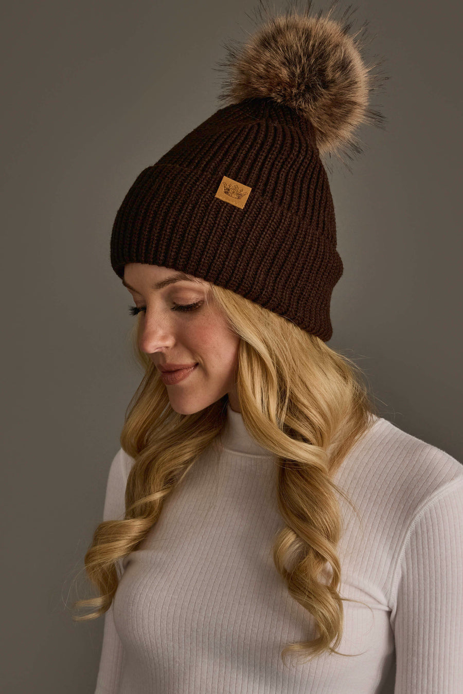 A woman with long, wavy blonde hair wears the Chocolate Brown Knit Pom Hat from Panache Apparel Collection, featuring a fur pom-pom, and a white ribbed turtleneck as she looks down against a neutral background.