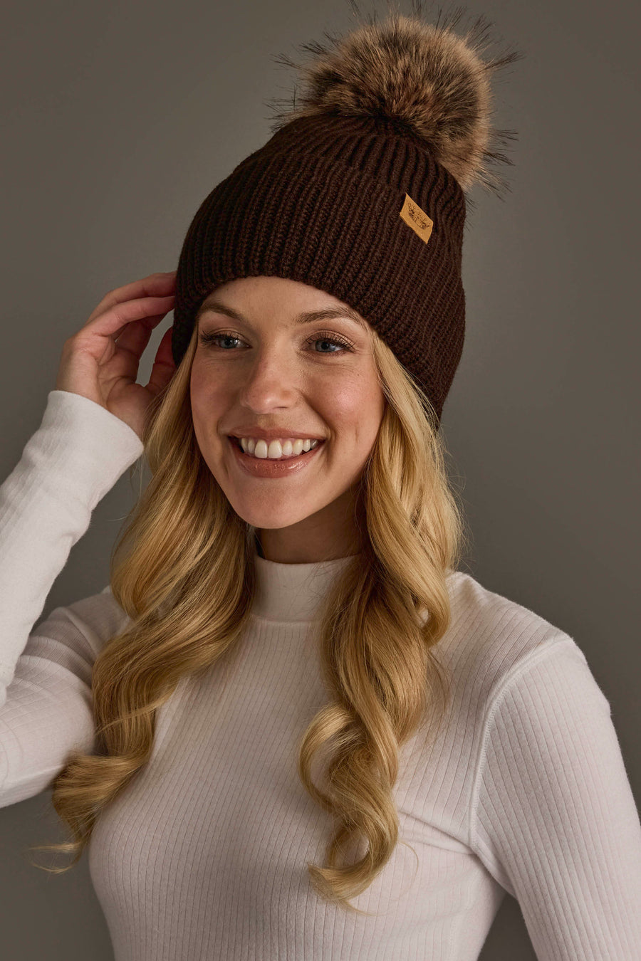 A smiling woman with long blonde hair models the Panache Apparel Collection’s Chocolate Brown Knit Pom Hat, featuring a fur pom-pom, along with a white ribbed long-sleeve shirt, posed against a plain gray background.
