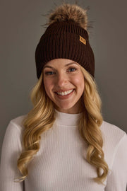 A smiling woman with long blonde hair wears the Panache Apparel Collection Chocolate Brown Knit Pom Hat and a white ribbed sweater, posing in front of a neutral background.