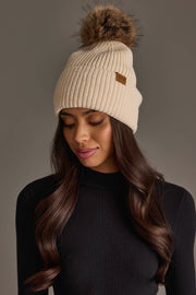 A woman models the Panache Apparel Collection’s Beige Knit Pom Hat, featuring a fur pom-pom, paired with a black ribbed long-sleeve top. She looks down with a soft smile against a plain gray background.