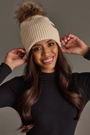 A woman smiles while wearing the Panache Apparel Collection Beige Knit Pom Hat, a cream-colored knit beanie with a fur pom-pom, paired with her long wavy dark hair and a black ribbed turtleneck sweater, standing against a plain grey background.