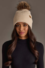 A woman with long, wavy hair wears the Beige Knit Pom Hat by Panache Apparel Collection and a black ribbed turtleneck sweater, standing against a plain gray background.