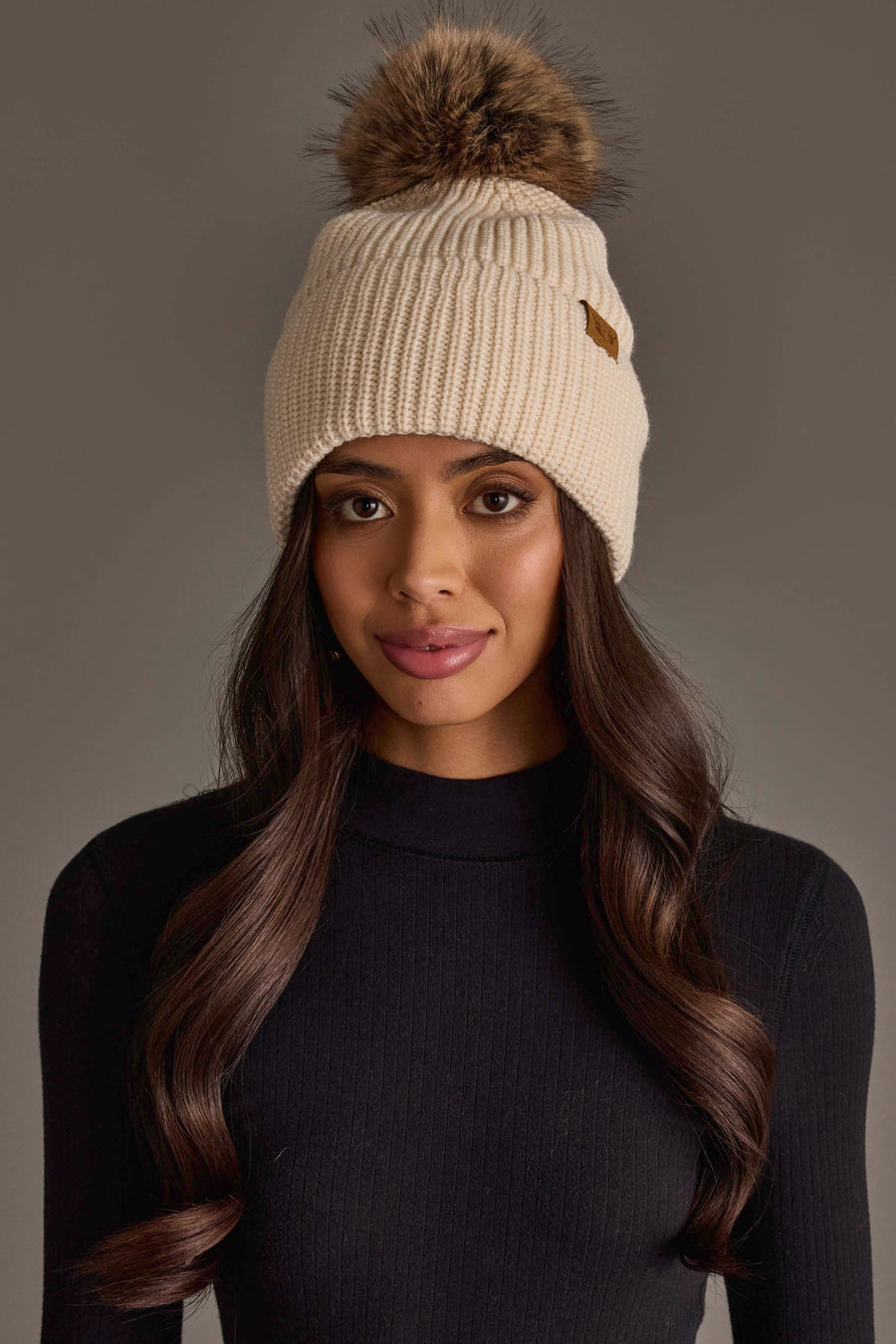 A woman with long, wavy hair wears the Beige Knit Pom Hat by Panache Apparel Collection and a black ribbed turtleneck sweater, standing against a plain gray background.
