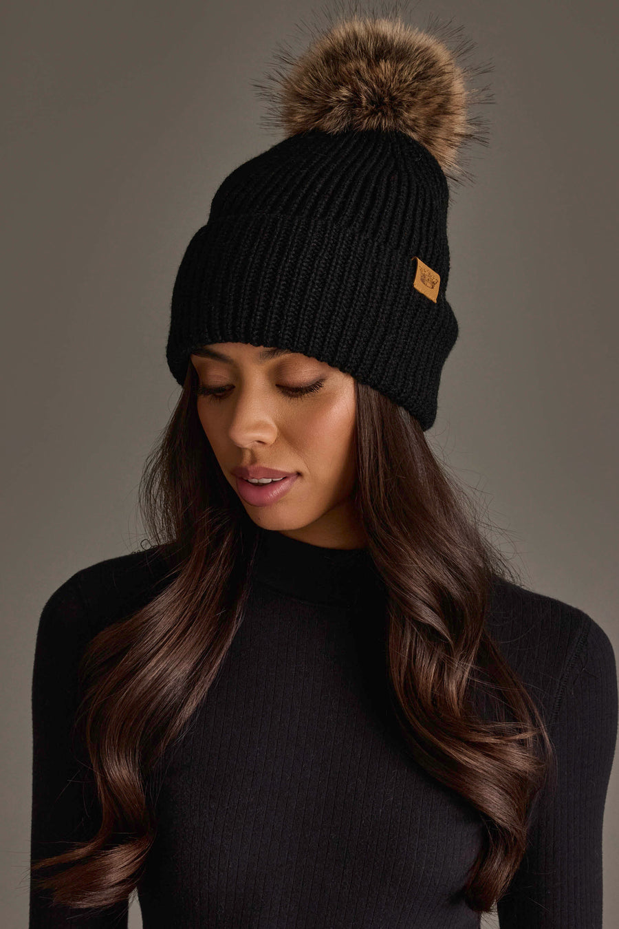 A woman with long brown hair wears the Panache Apparel Collection Black Knit Pom Hat, featuring a brown faux fur pom-pom, along with a black ribbed turtleneck sweater, looking down with a neutral expression.