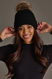 A smiling woman wearing the Panache Apparel Collection Black Knit Pom Hat, featuring a faux fur pom-pom, touches the sides of the hat. She has long, wavy hair and is dressed in a black long-sleeve top against a neutral background.