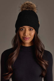 A woman with long wavy hair poses against a neutral background, wearing the Panache Apparel Collection Black Knit Pom Hat and a black ribbed sweater.