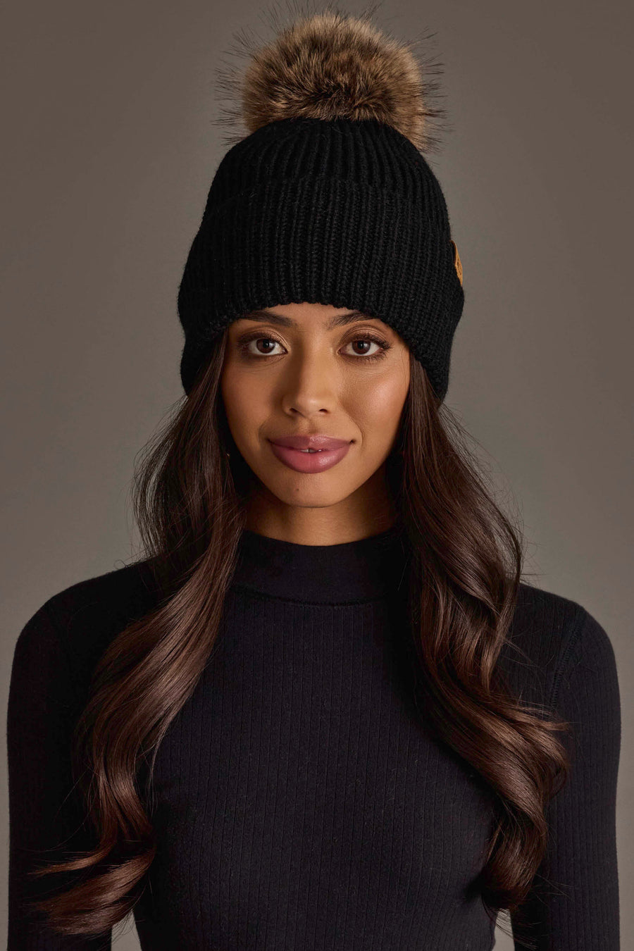 A woman with long wavy hair poses against a neutral background, wearing the Panache Apparel Collection Black Knit Pom Hat and a black ribbed sweater.
