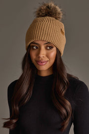 A woman with long brown hair is wearing the Panache Apparel Collection’s Tan Knit Pom Hat, featuring a faux fur pom-pom, along with a black ribbed sweater. She gazes slightly to the side against a plain gray background.