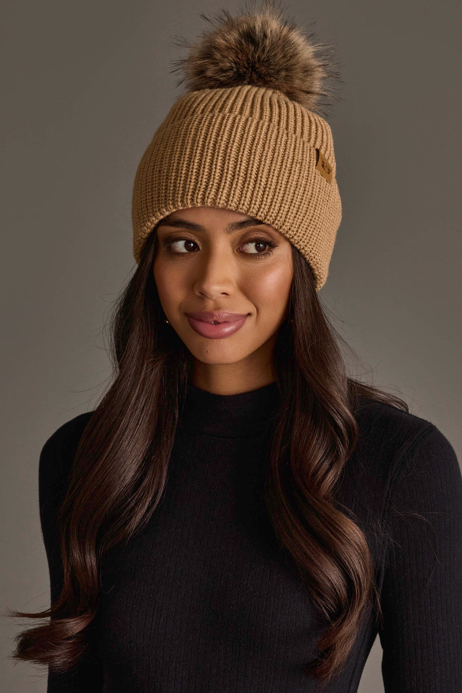 A woman with long brown hair is wearing the Panache Apparel Collection’s Tan Knit Pom Hat, featuring a faux fur pom-pom, along with a black ribbed sweater. She gazes slightly to the side against a plain gray background.