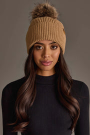 A woman with long, wavy brown hair wears the Panache Apparel Collection Tan Knit Pom Hat and a black ribbed sweater, looking directly at the camera against a plain gray background.
